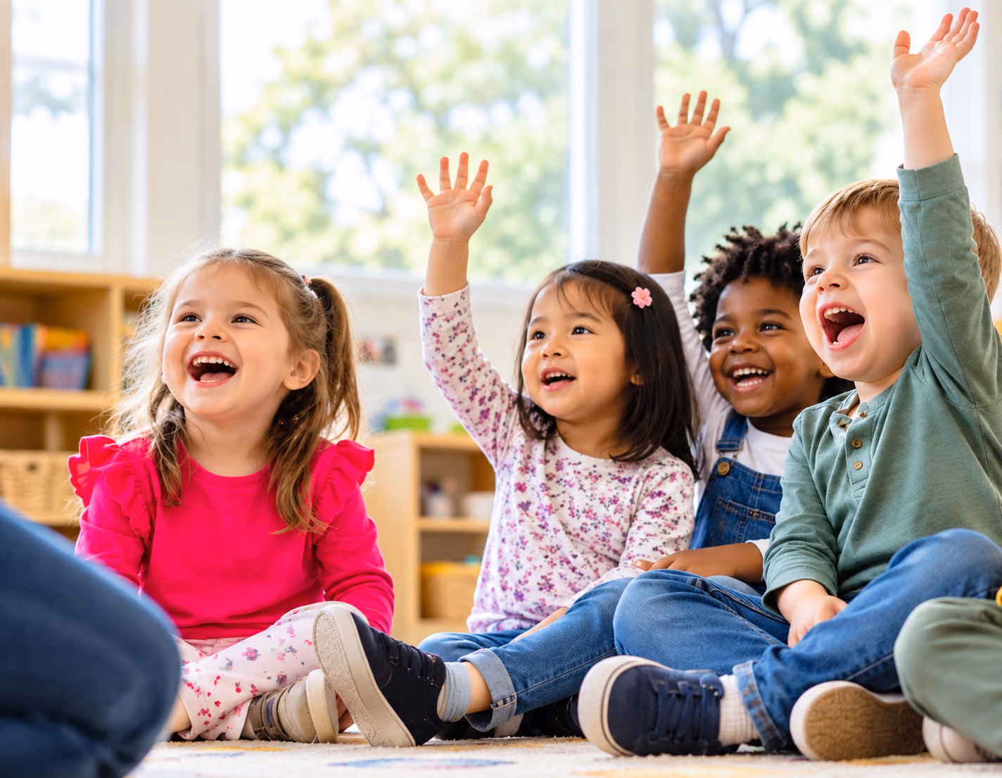 Children sitting together on the classroom floor, laughing with their hands raised