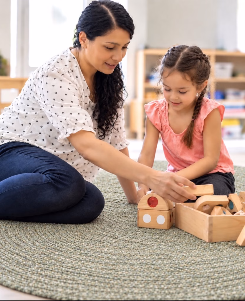 Teacher sitting on a classroom rug building with wooden blocks alongside a young girl